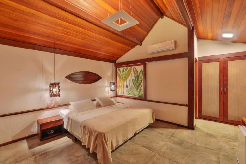Um quarto da Pousada Sitio da Prainha com uma cama king size, teto de madeira, duas mesas laterais, arte de parede com folhas e armário embutido.