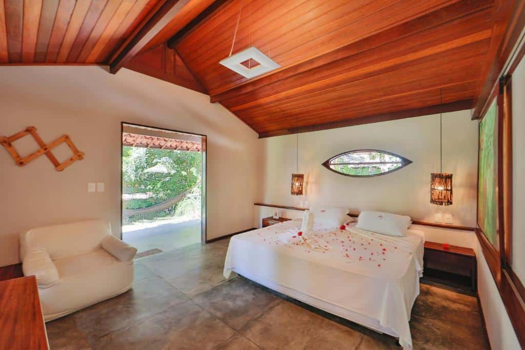 Um quarto da Pousada Sitio da Prainha com cama king-size, poltrona branca, teto de madeira e pétalas de rosa sobre a cama, com uma porta aberta para a vegetação. Essa é uma das indicações de onde ficar na Praia dos Carneiros.