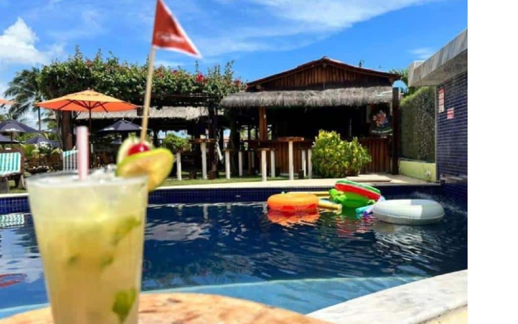 Um drinque tropical está em uma mesa em frente a uma piscina da Pousada Solar Dos Carneiros com bóias coloridas e um bar ao lado da piscina ao fundo.