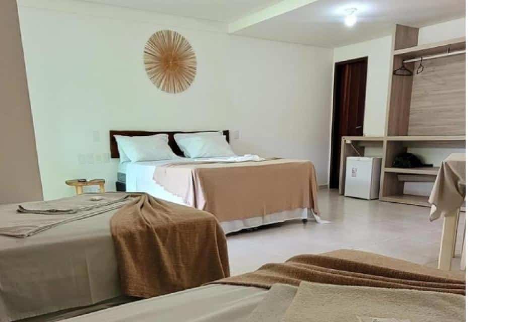 Um quarto da Pousada Solar Dos Carneiros com duas camas, roupa de cama bege, um armário aberto, um pequeno frigobar e decoração mínima na parede. Essa é uma das indicações de onde ficar na Praia dos Carneiros.