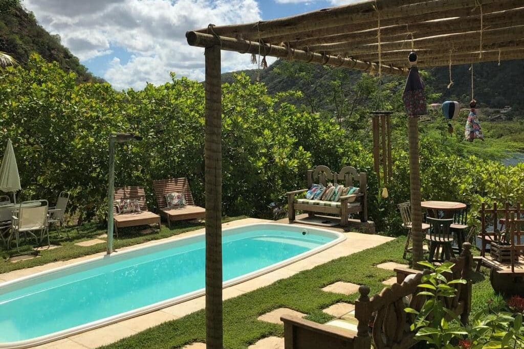Uma pequena piscina no quintal da Pousada Trilha do Velho Chico com bancos de madeira, cadeiras, mesas e uma pérgola, cercada por vegetação e colinas.
