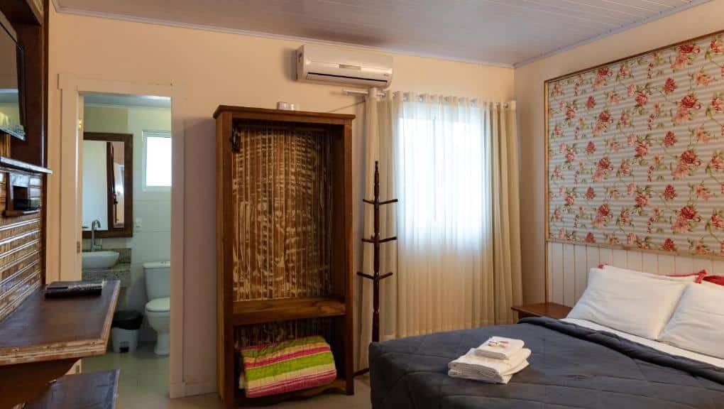 Quarto da Pousada UpSerra aconchegante com parede floral, móveis de madeira e cama confortável com cobertores escuros e toalhas.