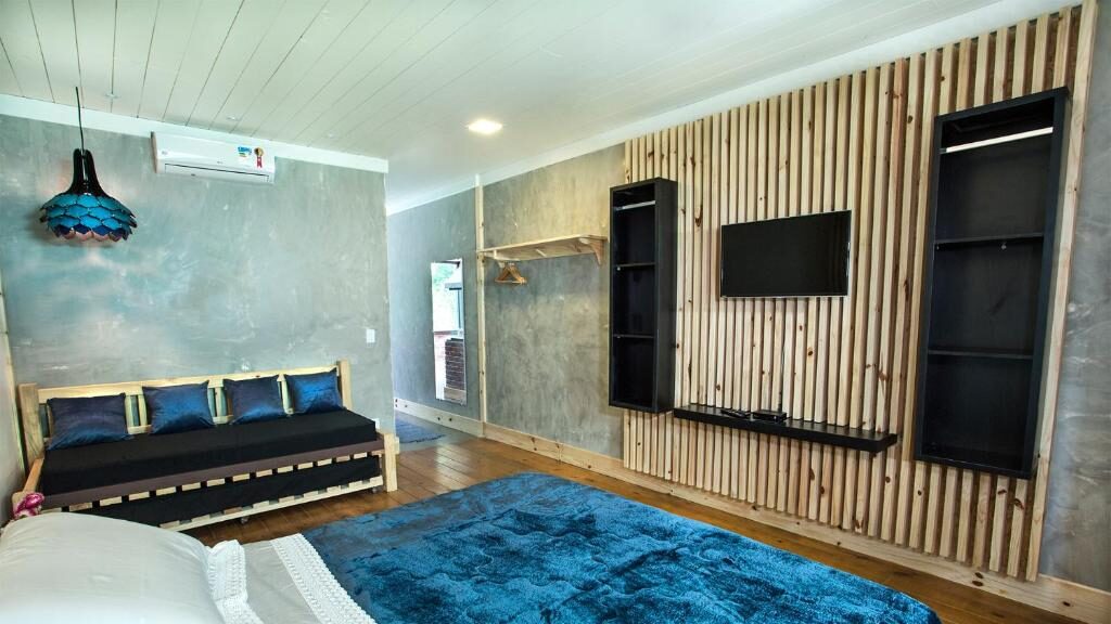 Um quarto da Pousada Valle do Avencal com uma colcha azul, um sofá-cama, uma TV na parede, prateleiras abertas e piso de madeira.