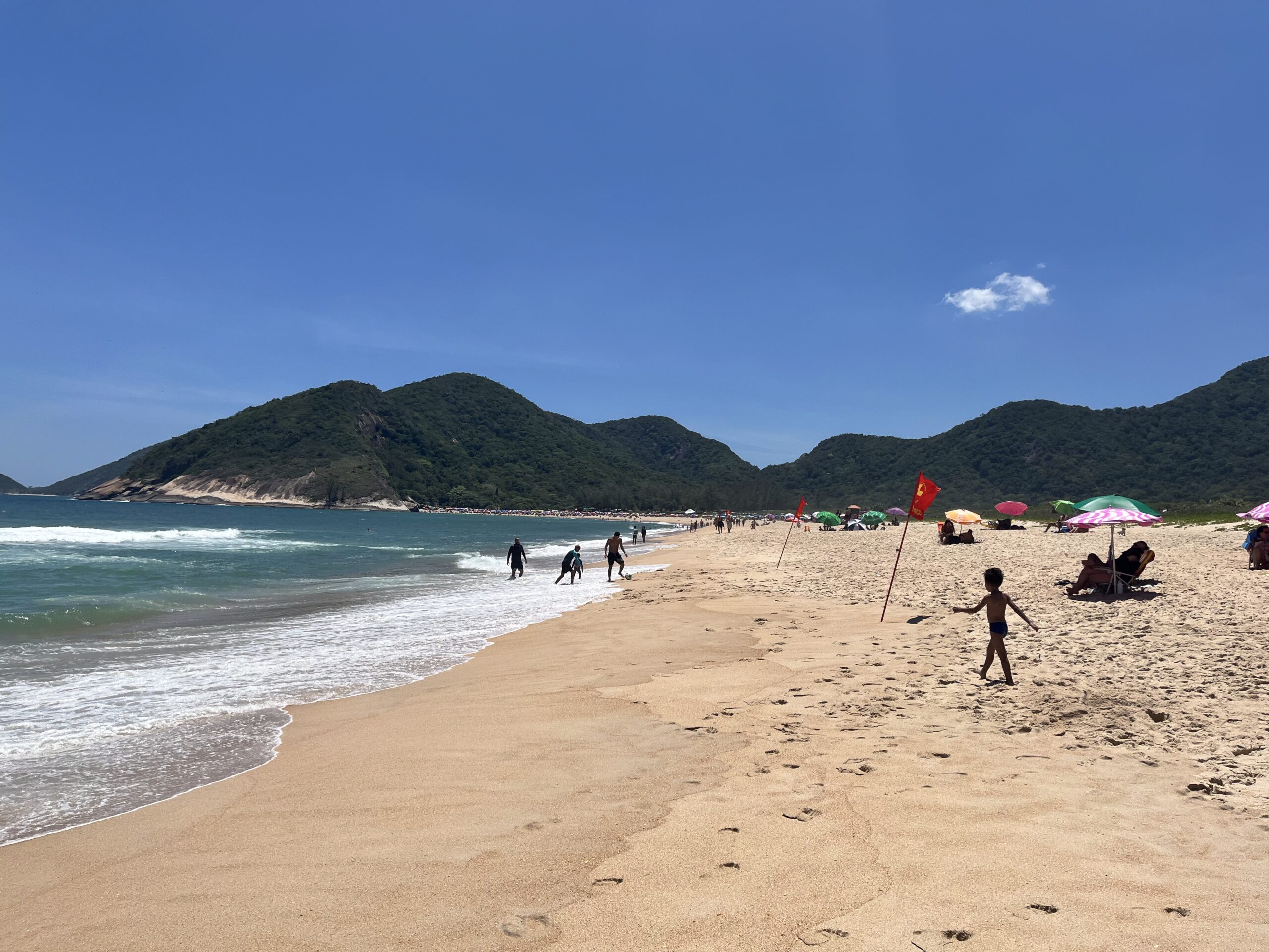 Uma praia movimentada com montanhas verdes ao fundo. Várias pessoas aproveitando o sol e as ondas na areia dourada. Crianças brincando no mar, guarda-sóis coloridos e barracas espalhadas pelo local.