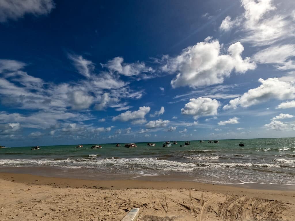 Praia de Maracajaú com areia com ondas suaves, vários barcos pequenos flutuando na água azul-turquesa e céu azul parcialmente nublado.