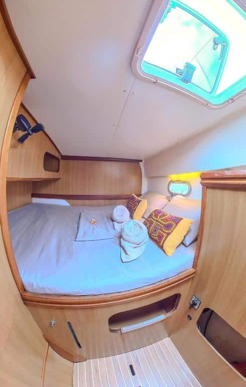 Uma cabine de barco no Enjoy Sailing Experience compacta com uma cama feita, duas toalhas enroladas, travesseiros amarelos, painéis de madeira e uma escotilha aberta no teto. É uma das opções de onde ficar em San Blas.
