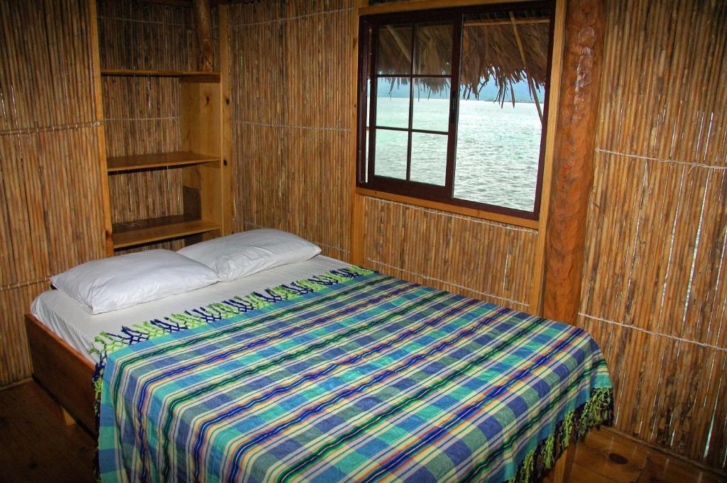 Uma cama pequena com um cobertor xadrez em um quarto da Private Cabin Over-the-Ocean including Meals com paredes de bambu, ao lado de uma janela com vista para a água. É uma das opções de onde ficar em San Blas.