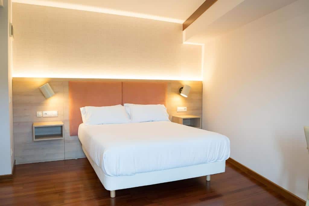 Uma cama bem arrumada do Hotel Parking Miradoiro de Belvís com roupas de cama brancas em um quarto moderno com piso de madeira e mesas de cabeceira embutidas.