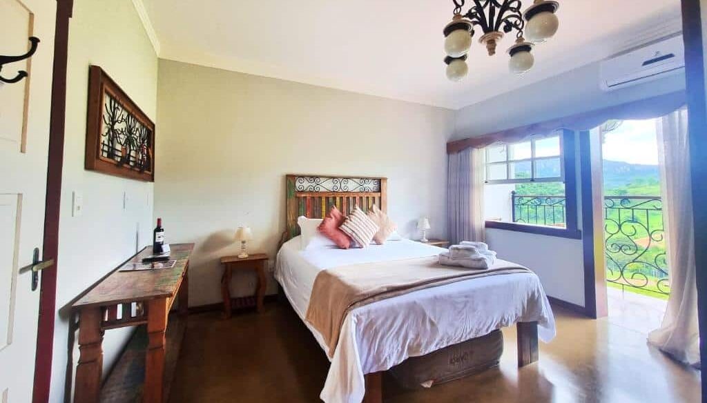 Uma cama bem arrumada em um quarto bem iluminado da Pousada Dom Xavier com uma escrivaninha de madeira, lustre e janela aberta com vista para a vegetação.