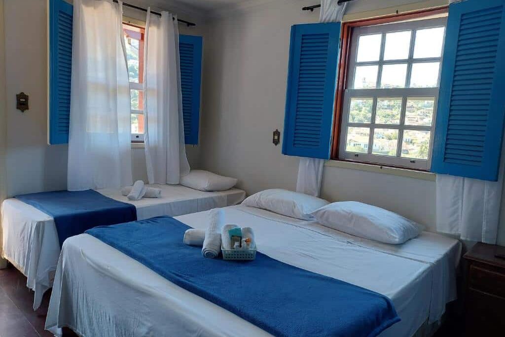 Um quarto bem iluminado da Pousada Ouro Preto com duas camas, cobertores azuis, lençóis brancos, persianas azuis e uma janela com vista para o exterior.