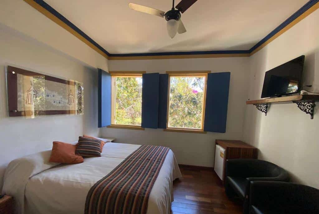 Um pequeno quarto da Pousada Relíquias do Tempo com uma cama, duas janelas, um ventilador de teto, uma TV em uma prateleira e duas cadeiras pretas.