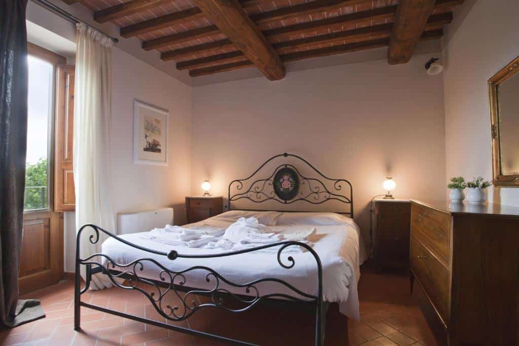 Um quarto do Relais Villa Belvedere com uma cama de ferro forjado, móveis de madeira, duas luminárias e uma janela aberta com cortinas que permitem a entrada de luz natural.