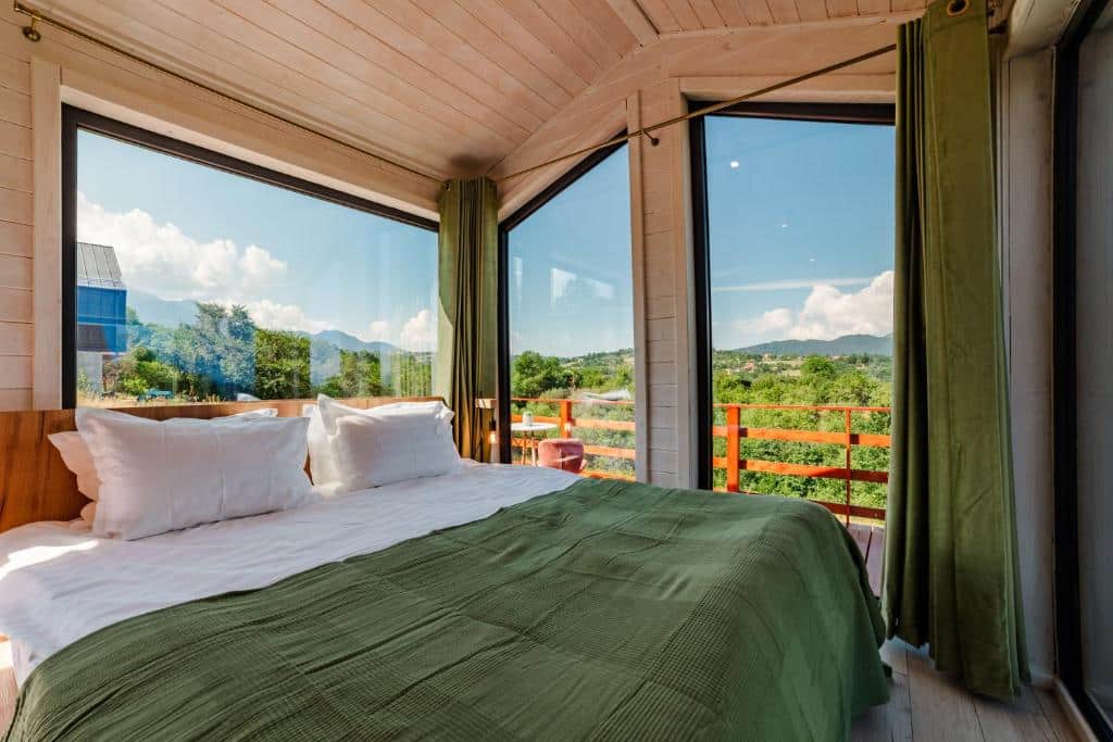 Um quarto do SkyAtlas Hideaway com uma cama grande, cobertor verde, travesseiros brancos e janelas do chão ao teto com vista para uma paisagem verde. Imagem para ilustrar post sobre hotéis na Romênia.