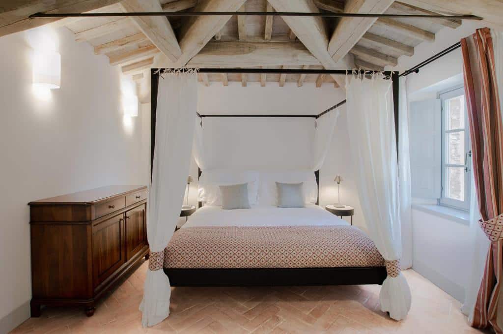 Quarto bem iluminado do Tenuta CastelGiocondo com uma cama de dossel, cortinas brancas, teto de madeira, cômoda e uma janela com cortinas estampadas.