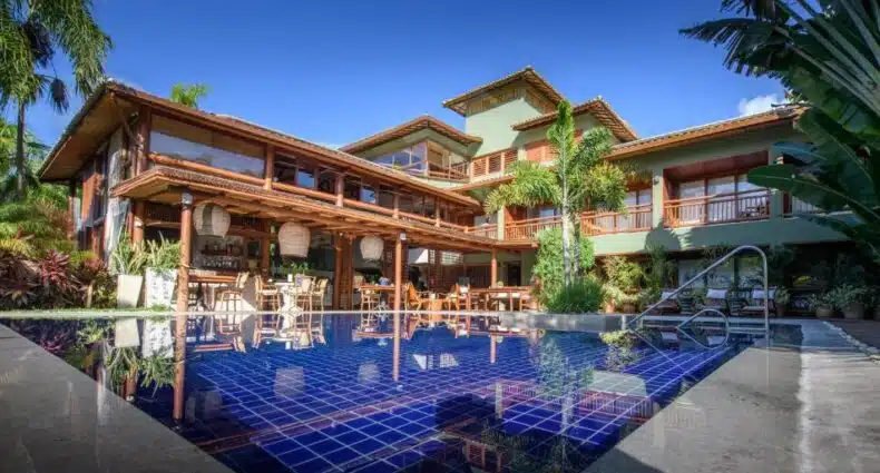 Casa grande de dois andares com detalhes em madeira, cercada por plantas e uma piscina de azulejos azuis em primeiro plano. Esse é o Refúgio da Vila Hotel Boutique & Spa, uma das dicas de onde ficar na Praia do Forte.