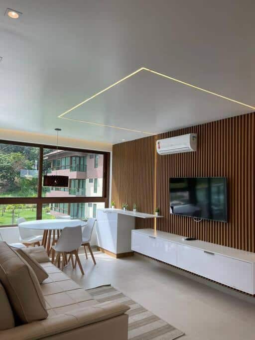 Moderna sala de estar e jantar na Reserva Carneiros beira-mar com painéis de madeira, uma TV na parede, móveis brancos e uma grande janela com vista para o exterior.