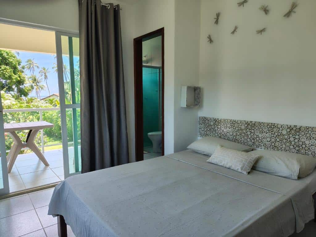 Um quarto do Sitio Recanto Feliz com uma cama bem arrumada, banheiro, arte na parede e uma varanda com uma mesa e vegetação do lado de fora. Essa é uma das indicações de onde ficar na Praia dos Carneiros.