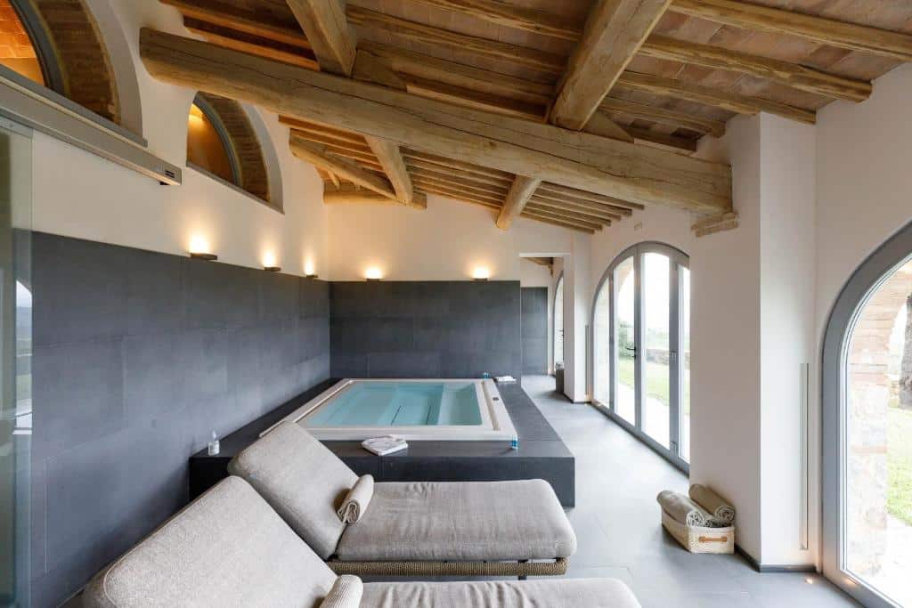 Sala de spa interna do Tenuta CastelGiocondo com duas espreguiçadeiras bege, uma banheira de hidromassagem quadrada, piso de azulejos, janelas grandes e vigas de madeira expostas. Imagem para ilustrar post sobre hotéis românticos na Toscana.