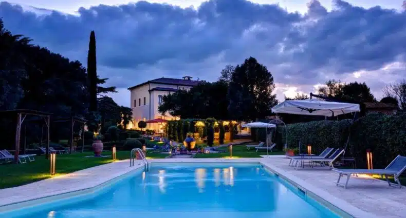 Uma piscina da Villa Sabolini ao anoitecer com espreguiçadeiras e uma vila iluminada ao fundo, cercada por árvores e arbustos. Imagem para ilustrar post sobre hotéis românticos na Toscana.