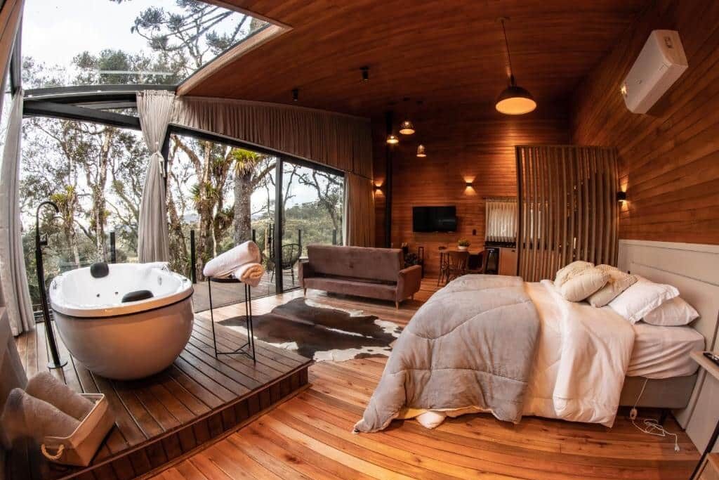 Um quarto moderno de madeira do Vale Tancon Eco Village Urubici com banheira redonda, cama, sofá, tapete de pele de vaca e janelas grandes com vista para as árvores.