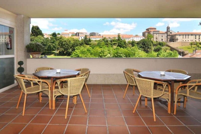 Pátio coberto do Hotel Parking Miradoiro de Belvís com mesas e cadeiras, com vista para uma paisagem verde e edifícios históricos à distância. Imagem para ilustrar post sobre hotéis em Santiago de Compostela.