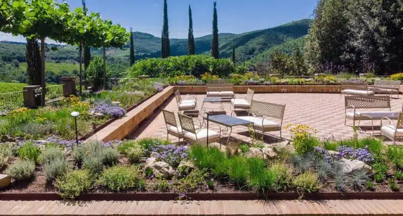 Pátio com cadeiras e mesas de metal no Villa Campomaggio Resort, cercado por jardins e árvores, com vista para as colinas verdes sob um céu azul. A foto é capa do post de onde ficar na Toscana.