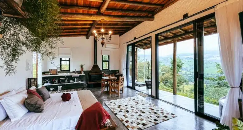 Aconchegante sala de estar com teto de madeira e janelas amplas com vista para a natureza verde ao redor. Piso de concreto polido, tapete de couro, mobília rústica e lareira de ferro criam um ambiente charmoso e convidativo, da Pousada Viver a Pedra, em Gonçalves. Representa pousadas no sul de Minas.