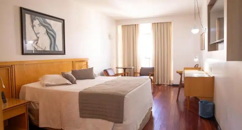 Quarto do Ritz Plaza Hotel com móveis de madeira, cama de casal, quadro decorativo na parede e janela com cortinas claras. Representa pousadas em Juiz de Fora.