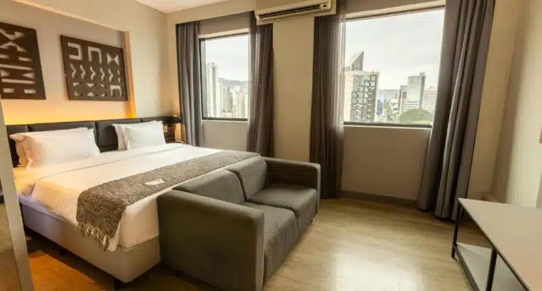 Quarto do Royal Golden Hotel - Savassi com vista panorâmica da cidade, decorado em tons neutros com móveis modernos e confortáveis. Na cama, há um edredom cinza e duas almofadas brancas. Junto à cama, há um sofá cinza. Grandes janelas oferecem uma vista impressionante da paisagem urbana. Representa hotéis na Savassi.