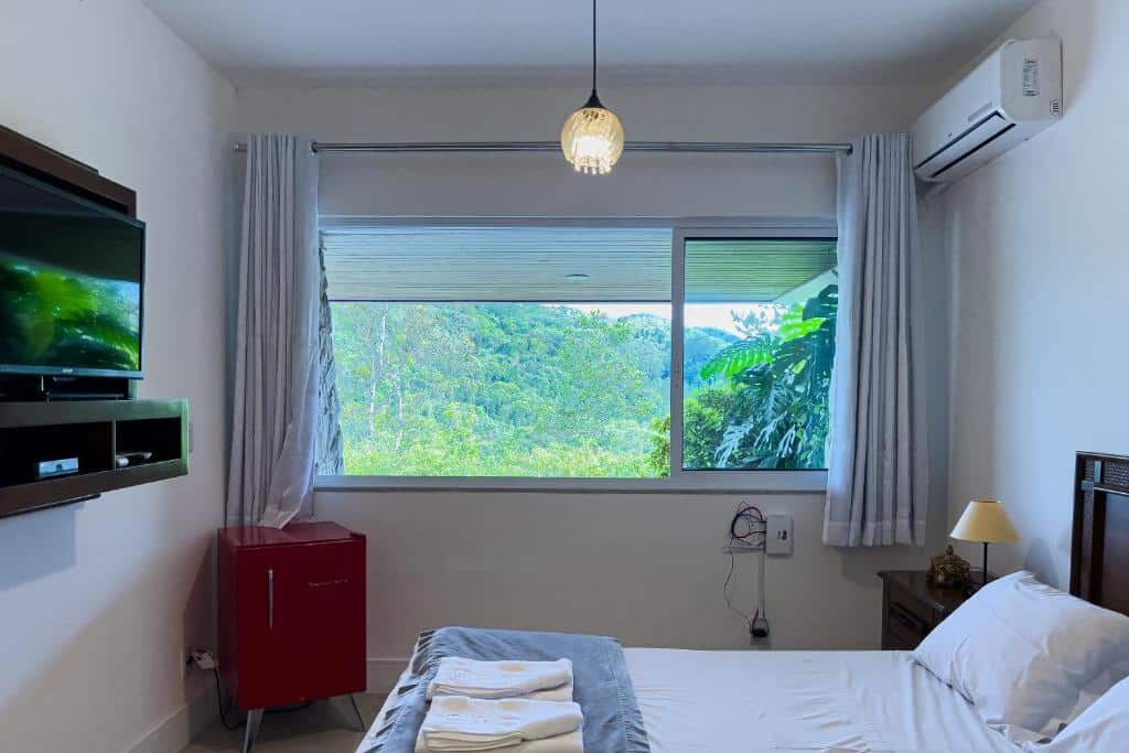 Quarto da Casa Branca Pousada com ampla janela panorâmica revelando bela paisagem natural com floresta verde e céu azul. A imagem mostra uma TV de tela plana, frigobar vermelho, abajur de mesa e cama com lençóis brancos e toalhas dobradas. Essa é uma das indicações de onde ficar em Pomerode.