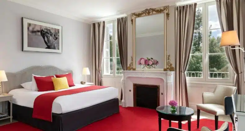 Quarto do Château Belmont elegante com cama de casal, espelho dourado sobre lareira e cortinas luxuosas. Decoração sóbria com tons neutros e toques de vermelho nos adornos. Imagem para ilustrar post sobre onde ficar no Vale do Loire.