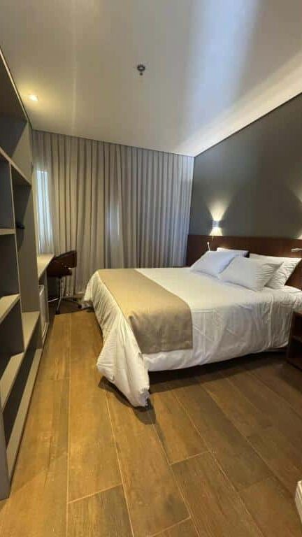 Quarto do Hotel C Jaguariúna com cama de casal, móveis de madeira escura e cortinas claras.