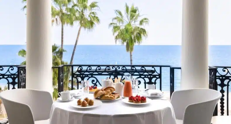 Vista de um restaurante do Hotel Le Negresco com vista deslumbrante para o mar e palmeiras ao fundo. Sobre a mesa há uma variedade de itens para um café da manhã: pães, frutas, suco e bebidas quentes em xícaras brancas. A mesa está decorada com toalha branca e há cadeiras confortáveis ao redor. Imagem para ilustrar post sobre onde ficar em Nice.