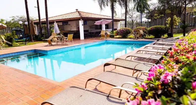 Piscina do Hotel Residence Jaguary cercada por palmeiras e floreiras com flores coloridas, com construção de estilo colonial e deck com espreguiçadeiras à beira d'água. Essa é uma das indicações de onde ficar em Jaguariúna.