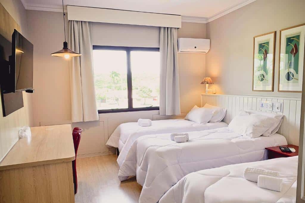 Quarto do Hotel Residence Jaguary aconchegante com duas camas de casal, decoração neutra e acessórios, janela com vista para área externa verde.
