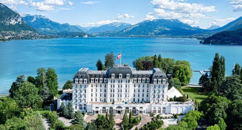Imponente hotel histórico com arquitetura neoclássica às margens de lago montanhoso cercado por belas paisagens naturais, com bandeira suíça esvoaçando no alto.