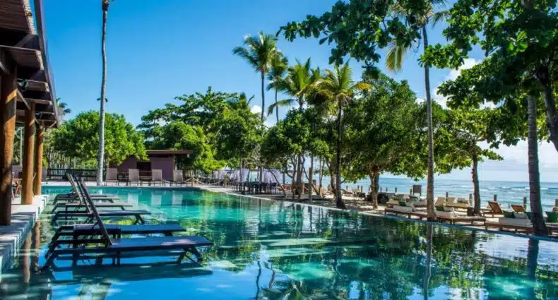 Piscina do Kûara Hotel com espreguiçadeiras e palmeiras refletida na água cristalina sob céu azul. Imagem para ilustrar post sobre onde ficar em Arraial d'Ajuda.