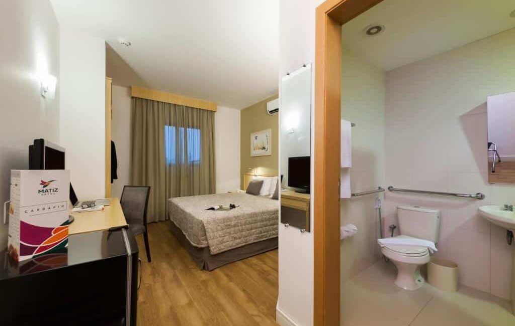 Quarto do Matiz Jaguariúna com cama de casal, mesinha com computador portátil, e banheiro com pia e vaso sanitário.