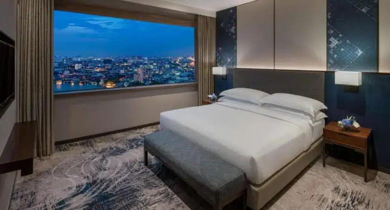 Imponente quarto do Millennium Hilton Bangkok com vista panorâmica da cidade iluminada à noite, com arranha-céus e luzes brilhantes refletidas no céu azul escuro estrelado. Imagem para ilustrar post sobre onde ficar em Bangkok.