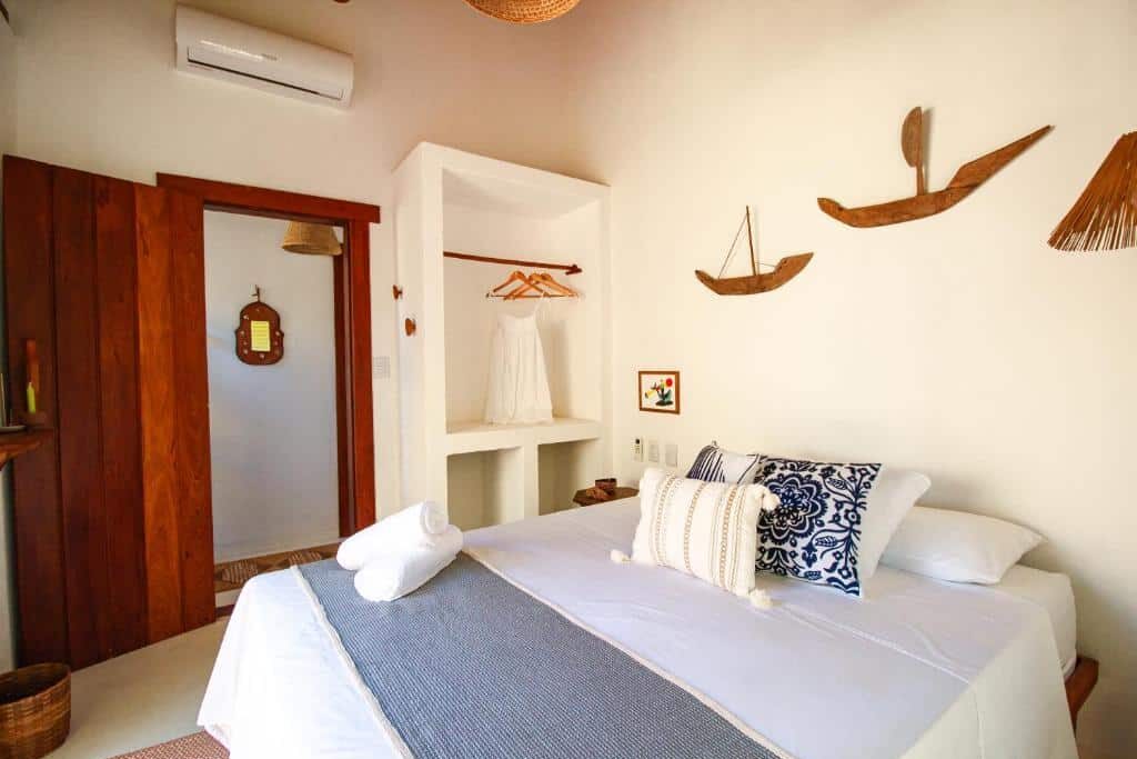 Quarto da Morada Pochixá com decoração leve e rústica, cama de casal com roupas de cama brancas, prateleiras e adornos de madeira na parede, ventilador de teto e ar-condicionado instalado. Essa é uma das dicas de onde ficar em Corumbau.