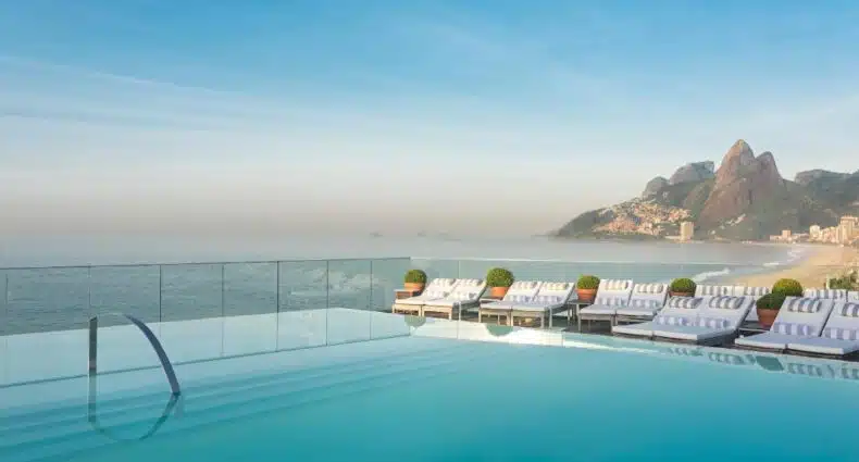 Vista panorâmica da praia de Ipanema com piscina e espreguiçadeiras à beira-mar, com a Pedra da Gávea ao fundo em um dia ensolarado, para representar onde ficar em Ipanema.