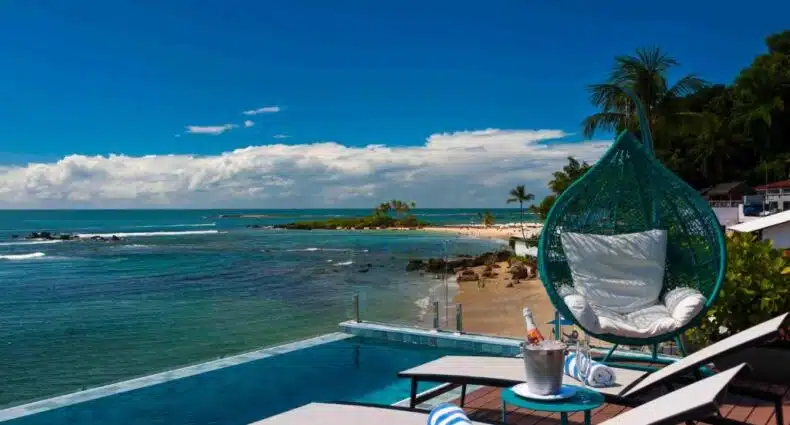 Vista panorâmica do litoral paradisíaco da Bahia com praia de areia clara, águas azul-esmeralda e coqueiros, vista de rede suspensa em área externa de resort de luxo. Representa Pousada Antonella, na Bahia