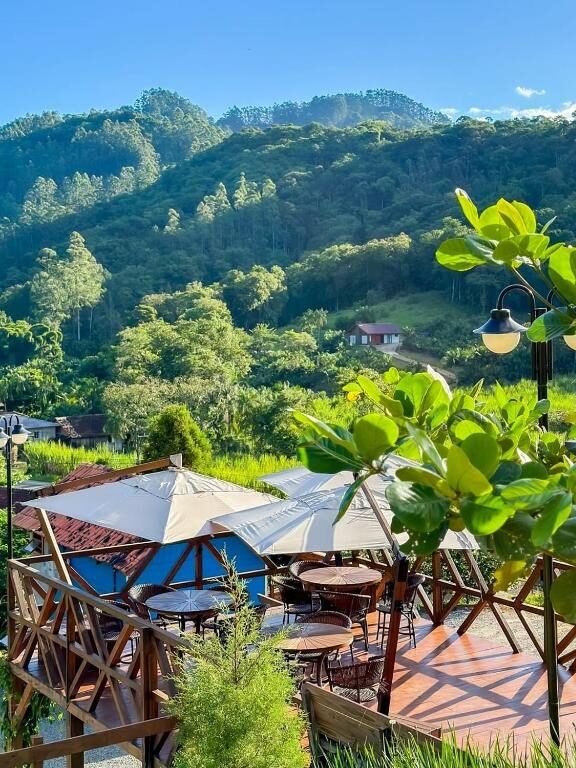 Cenário idílico em área rural da Pousada Hoff Haus com chalés, mesas e guarda-sóis sob bela vegetação.