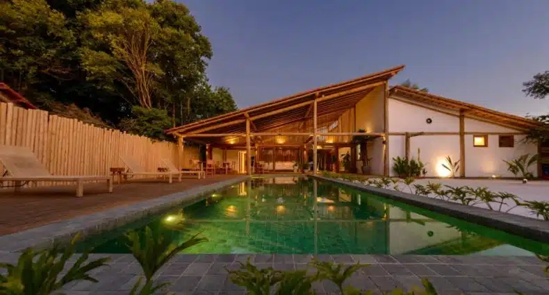 Pousada Nossa Casa Cumuruxatiba com telhado de madeira, piscina e vegetação ao redor durante a noite iluminada. Essa é uma das dicas de onde ficar em Cumuruxatiba.