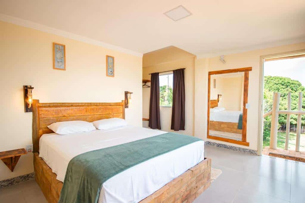 Quarto da Pousada Vila do Sol com decoração rústica e acolhedora, contendo cama de madeira, cortinas escuras, espelho de parede e vista externa com natureza. Essa é uma das dicas de onde ficar em Corumbau.