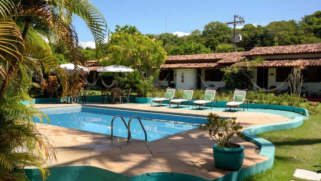 Piscina cercada por vegetação exuberante na Pousada Villa Cumuru, com espreguiçadeiras verdes convidando ao descanso