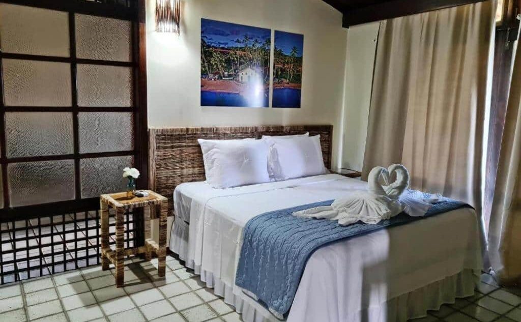 Quarto aconchegante do Hotel Praia dos Carneiros com cama de casal, criado-mudo de palha e decoração tropical em parede com paisagem litorânea. É uma das opções de onde ficar em Tamandaré.