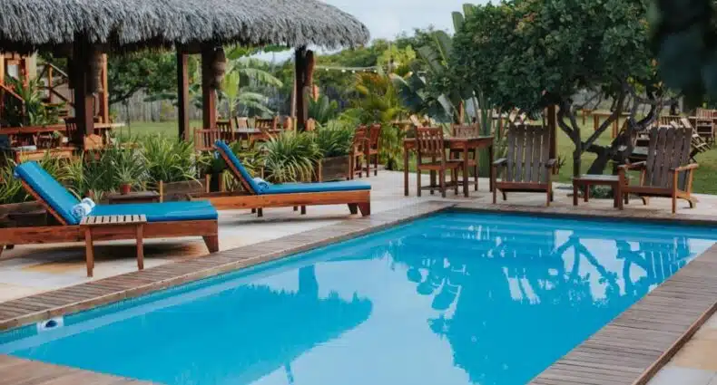 Piscina da Vila Aty Lodge cercada por cabanasde palha em complexo de resort tropical com espreguiçadeiras azuis à beira d'água e vegetação exuberante ao redor. Imagem para ilustrar post sobre onde ficar nos Lençóis Maranhenses.