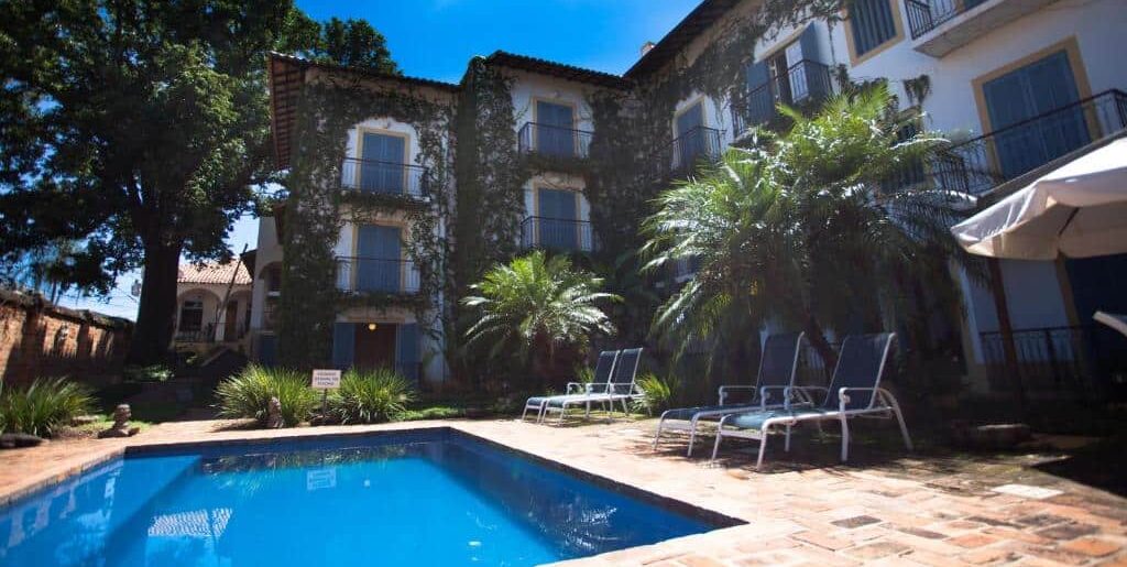 Um pátio da Vila Bueno Residence com piscina e cadeiras de praia, cercado por palmeiras e árvores em um belo cenário tropical. Essa é uma das dicas de onde ficar em Jaguariúna.
