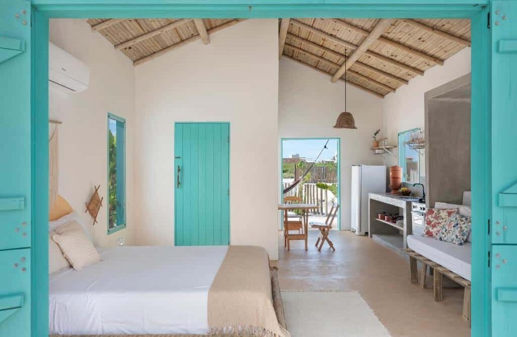 Quarto acolhedor em estilo rústico na Vila da Falésia com teto de madeira e paredes claras, porta e janela em tom de turquesa contrastando com a decoração neutra. Cama com roupa de cama leve e mesa de madeira com geladeira e utensílios domésticos. Essa é uma das dicas de onde ficar em Corumbau.